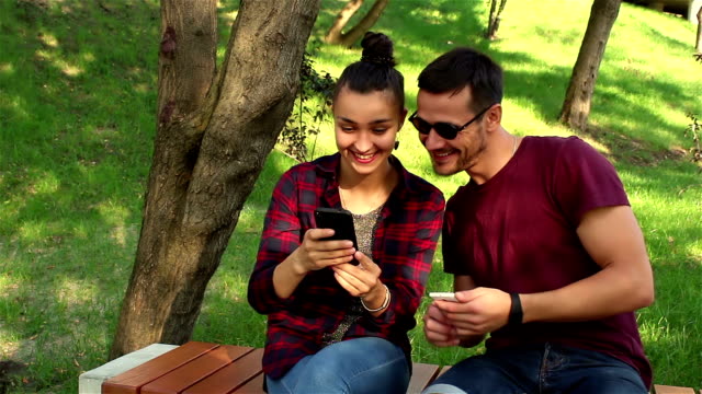 Couple-in-love-show-photos-on-their-smartphones-with-each-other,-sitting-on-a-park-bench.