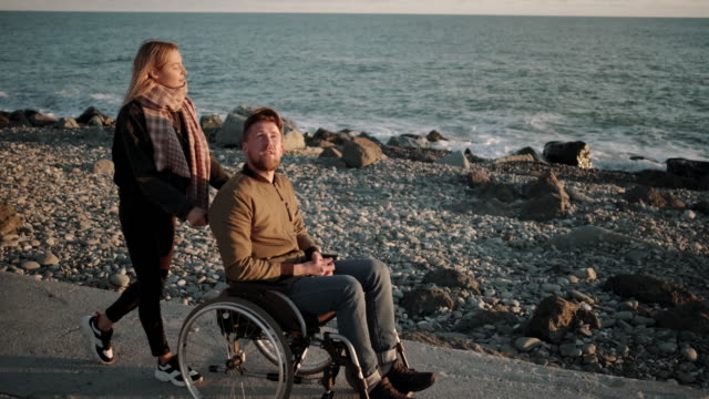 Wife-is-rolling-carriage-of-her-disabled-husband-over-embankment-with-sea-view