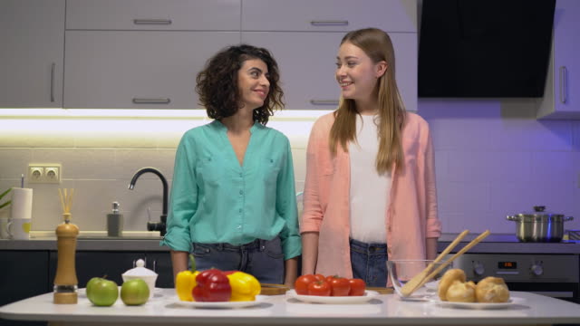 Two-young-friends-smiling-on-cam,-looking-at-each-other,-cooking-in-the-kitchen