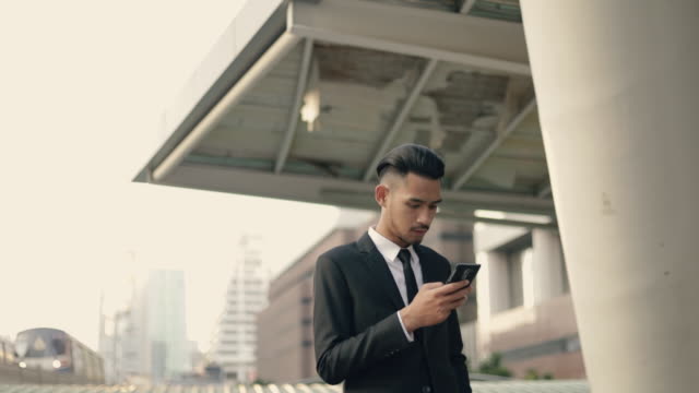 Guapo-joven-hombre-de-negocios-usando-un-teléfono-inteligente-enviando-mensajes-de-correo-electrónico-en-línea-en-la-calle-urbana-que-tiene-sky-train-en-el-fondo.