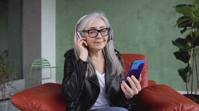 Front-view-of-beautiful-smiling-modern-60-aged-woman-with-long-gray-hair-which-wearing-glasses-and-enjoying-nice-music-on-headphones