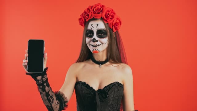 Día-de-Muertos,-hembra-con-maquillaje-cráneo-levantando-el-teléfono-celular,-apuntando-a-su-pantalla-por-dedo-índice-y-sonriendo-posando-sobre-fondo-rojo,-Halloween
