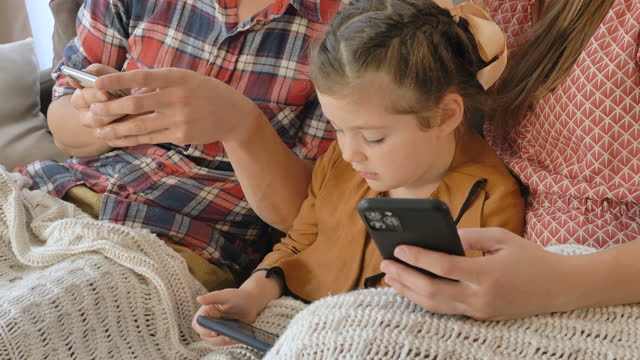 Little-girl-with-parents-plays-in-phones-sitting-on-sofa