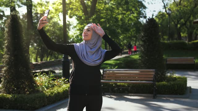 Attraktive-muslimische-Dame-in-lässigen-Kleidern-und-grauem-Hijab.-Sie-lächelt-und-macht-Selfie-auf-ihrem-Smartphone,-während-sie-in-einem-grünen-Landschaftspark-posiert
