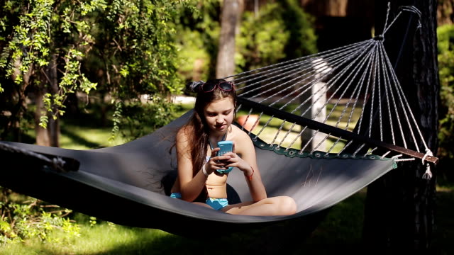 Eine-Mädchen-in-einem-blauen-Badeanzug-nutzt-ihr-Handy-und-Schaukeln-in-einer-Hängematte-in-Natur.