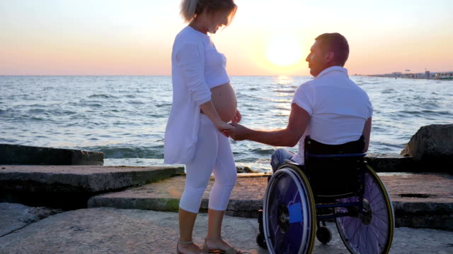 Handicapped-with-pregnant-girl-watching-sunset,-spouse-in-wheelchair-holds-arm-woman-with-big-belly,-happy-couple