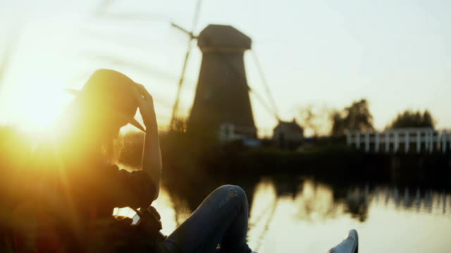 Millennial-girl-sits-with-phone-on-lake-pier.-Setting-sun.-Woman-using-shopping-app-outdoors.-Old-Dutch-windmill.-4K