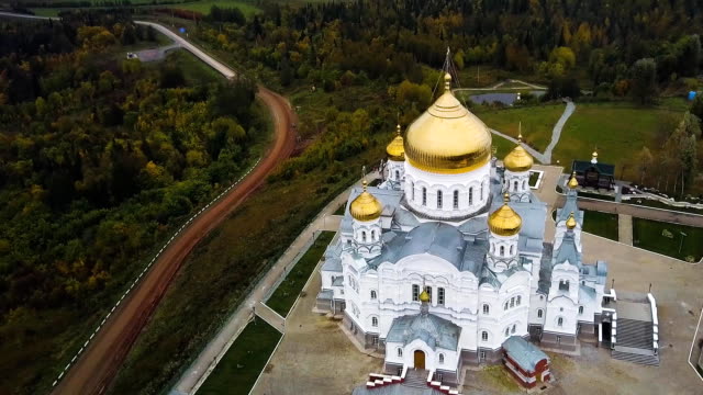 Aerial-view-on-great-white-christian-church-with-golden-domes-on-the-hill-and-field-with-forest-background.-Clip.-Top-view-on-Temple-in-the-middle-of-the-forest