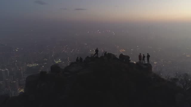Silhouettes-of-people-on-the-top-of-rock-at-twilight.-Illuminated-City-Skyline-in-Smog.-Aerial-View.-Drone-is-Flying-Sideways.-Establishing-shot.