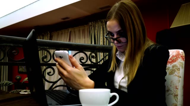 Business-Mädchen-mit-Brille-und-strengen-Anzug-Smartphone-vor-Laptop-benutzen