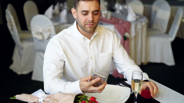 Loving-boyfriend-is-waiting-for-his-girl-in-restaurant,-using-smartphone,-opening-jewelry-box-and-looking-at-ring.-Bouquet-of-roses-and-glass-of-champagne-are-visible.