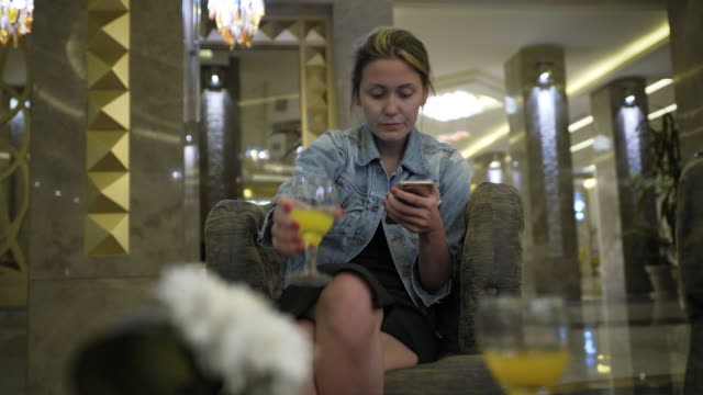 young-Caucasian-woman-in-a-jeans-jacket-drinks-juice-from-a-glass-and-uses-a-smartphone,-writes-messages-on-the-social-network.-Sits-at-the-table-in-the-hotel-restaurant.-Concept-of-healthy-fresh-food-in-business