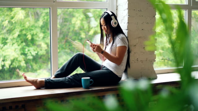 Fröhlich-Asiatin-Lieblingssong-im-Kopfhörer-hört-zu-und-mittels-Smartphone-Lächeln-und-Lachen-auf-der-Fensterbank-sitzen.-Internet-und-Jugend-Kultur-Konzept.