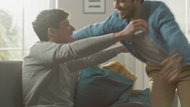 Sweet-Male-Gay-Couple-Full-Around-on-a-Sofa-at-Home.-Boyfriend-Runs-and-Jumps-into-Hands-of-His-Partner.-They-Hug.-They-are-Happy-and-Laughing.-They-are-Casually-Dressed-and-Room-Has-Modern-Interior.