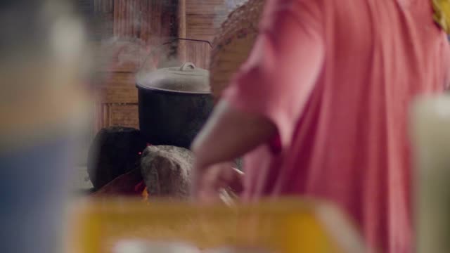 Mujer-soplando-fuego-con-ventilador-mientras-cocina-comida-en-fogata-en-maceta-en-la-aldea-asiática.-Mujer-cocinando-la-cena-tradicional-en-caldero-en-leña-ardiente-en-el-campo.