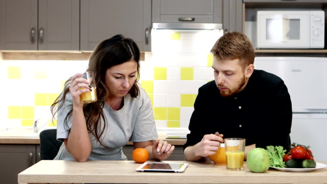 Young-couple-man-and-woman-with-tablet-eating-breakfast-sitting-by-table-in-kitchen-at-home.
