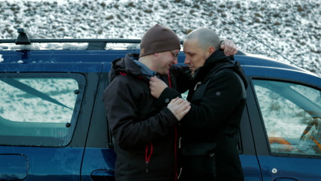Two-LGBT-guys,-bald-with-a-beard-and-wearing-glasses-and-a-hat-hugging-and-chatting-on-the-side-of-the-road-near-the-car