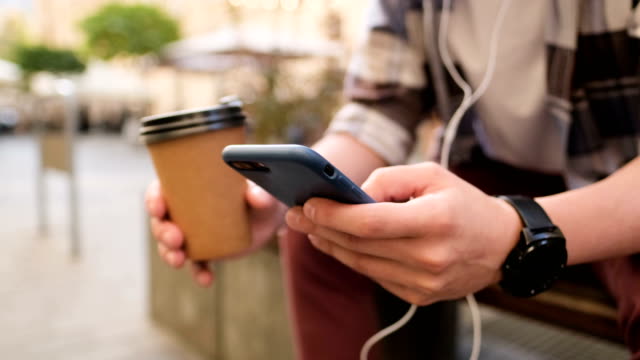Mann-in-Kopfhörern-mit-Smartphone,-Musik-hören-und-auf-Smartphone-surfen.-Kaffee-trinken,-um-ins-Freie-zu-gehen.-Nahaufnahme.-Blogger-scrollt-durch-soziale-Medien-auf-Gerät,-lesen-Nachrichten-auf-App