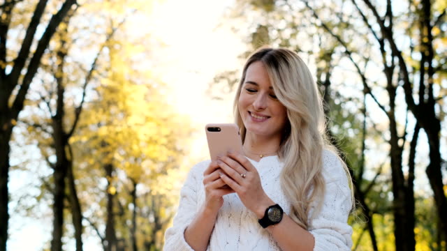 Portrait-of-girl-in-white-sweater-writing-message-on-her-smartphone-outdoors.-Woman-using-digital-gadget,-scrolls-through-social-media-on-device,-reading-news-on-app,-at-sunset