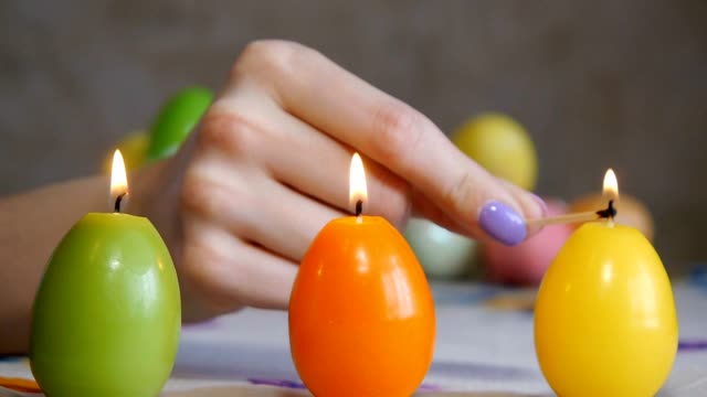 Candles-made-in-shape-of-easter-egg.-Green,-orange,-yellow.-Female-hand-lights-yellow-candles.-Two-candles-are-burning-nearby.