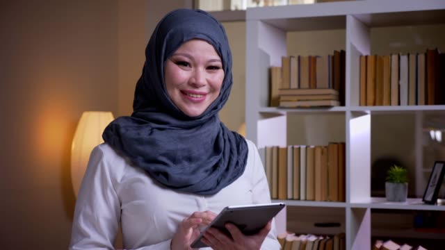 Tiro-de-primer-plano-de-la-empleada-femenina-musulmana-adulta-en-hijab-usando-la-tableta-mirando-a-la-cámara-y-sonriendo-felizmente-en-el-lugar-de-trabajo-en-el-interior