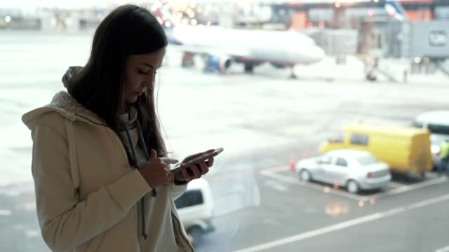 Attractive-girl-standing-at-window-in-airport-terminal-and-using-phone-in-background-of-airplane