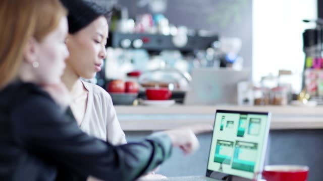 Zoom-out-of-two-young-businesswomen-discussing-website-templates-presented-on-laptop-screen-sitting-at-table-in-cafe,-Asian-woman-explaining-design-to-Caucasian-colleague