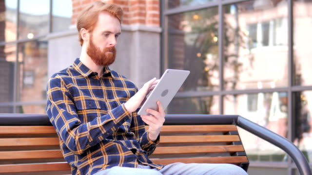 Barba-pelirroja-Joven-hombre-usando-la-tableta-mientras-está-sentado-en-el-banco