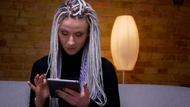 Closeup-shoot-of-young-attractive-caucasian-hipster-female-using-the-tablet-and-looking-at-camera-sitting-on-the-couch-indoors-in-a-cozy-apartment