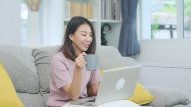 Junge-Geschäftsfreie-asiatische-Frau-arbeiten-auf-Laptop-Check-Social-Media-und-Kaffee-trinken,-während-auf-dem-Sofa-liegen,-wenn-im-Wohnzimmer-zu-Hause-entspannen.-Lifestyle-Frauen-im-HausKonzept.
