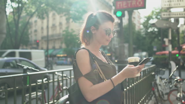 Junge-attraktive-Frau-schreibt-eine-SMS-auf-ihrem-Smartphone-an-der-U-Bahn-Ausfahrt-in-der-Straße,-trinken-ihren-Kaffee,-während-sonnigen-Sommer-in-Paris.-Sommersprossen,-Sonnenbrille,-Piercings,-rote-Haare,-4K-UHD.