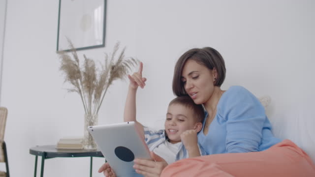 Mutter-und-Sohn-schauen-auf-den-Tablet-Bildschirm,-der-auf-einem-weißen-Bett-liegt.-Spiele-mit-Ihrem-Sohn-auf-Ihrem-Tablet-Computer-und-sehen-Sie-lustige-Videos