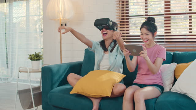 Joven-pareja-lesbiana-asiática-usando-tableta-y-realidad-virtual-jugando-juegos-juntos-mientras-se-acuesta-sofá-en-casa.