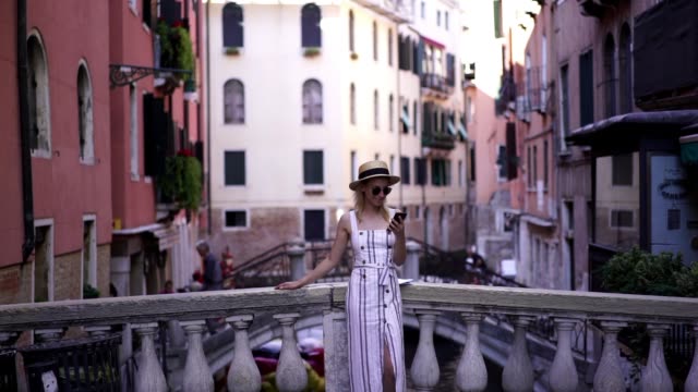 Slow-motion-effect-of-cheerful-hipster-girl-dressed-in-casual-look-resting-on-Venice-bridge-with-smartphone-gadget-in-hands