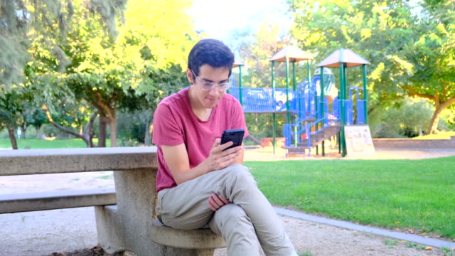 Young-man-using-his-smart-phone-sitting-in-a-park-bench