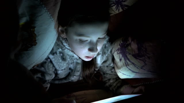 Ein-Kind,-ein-kleines-Mädchen,-versteckte-sich-nachts-unter-einer-Decke-auf-dem-Bett-und-spielt-auf-einem-Tablet-im-Dunkeln.-Konzeptvideo.-Internetsucht.-Nahaufnahme-des-Gesichts.-Rohes-Video.-4K.