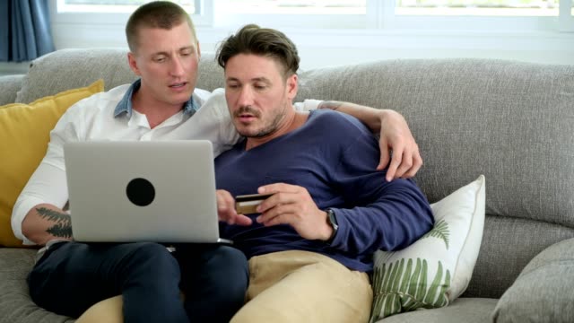 Pareja-gay-relajándose-en-el-sofá-usando-la-computadora-portátil.-Introducción-al-número-de-tarjeta-de-crédito.