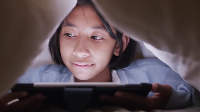 Asiática-niña-usando-tableta-bajo-manta-en-el-dormitorio-por-la-noche