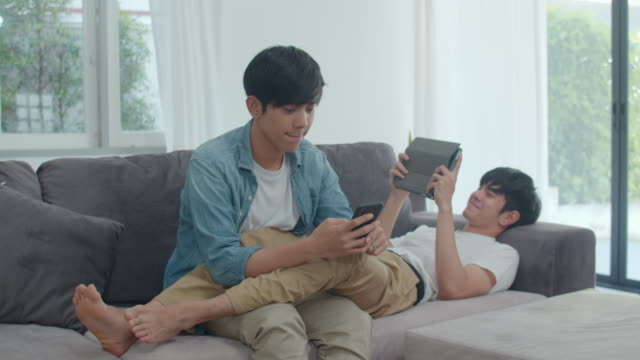 Young-gay-lgbtq-couple-using-mobile-phone-and-tablet-at-modern-home.-Asian-lover-male-happy-relax-laugh-and-fun-technology-play-games-in-internet-together-while-lying-sofa-in-living-room-concept.