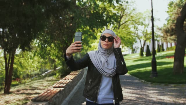 Muslim-female-in-casual-clothes,-sunglasses-and-hijab.-She-is-smiling,-enjoying-online-video-call-on-her-smartphone-while-walking-in-park.-Сlose-up