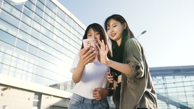 Schöne-lächelnde-asiatische-Mädchen-mit-langen-Haaren-und-in-stilvollen-Kleidern-gekleidet,-die-zusammen-in-der-Nähe-des-Flughafens-stehen-und-ihr-Telefon-benutzen