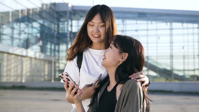 Zwei-schöne-ausgelassene-junge-asiatische-Mädchen-lächeln-aus-Revision-von-lustigen-Fotos-auf-dem-Handy-in-der-Nähe-von-modernen-Gebäude