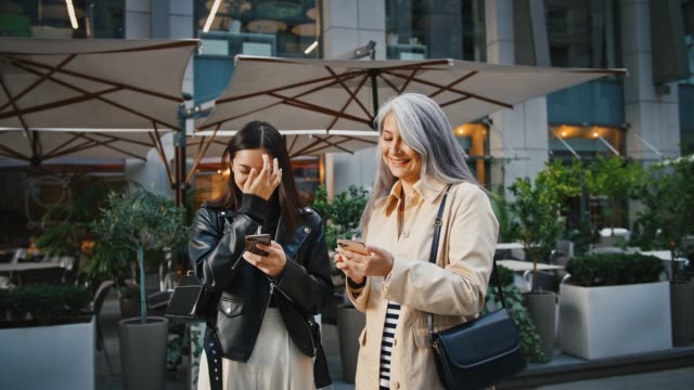 Asian-mature-mom-and-adult-daughter-are-using-their-smartphones-and-laughing-while-standing-near-terrace-of-restaurant