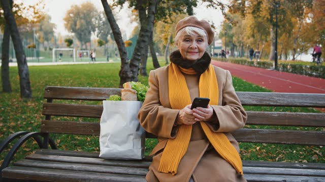 Elderly-lady-using-smartphone,-taking-deep-breath-sitting-on-bench-in-autumn-park-next-to-paper-bag-of-groceries