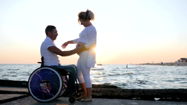 Pregnant-female-at-sunset-walks-up-to-disabled-man-in-a-wheelchair.-The-man-caresses-the-womans-pregnant-stomach-on-embankment