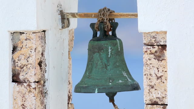 Old-rusty-bell-hanging-in-white-arch-against-blue-background,-church-in-Greece