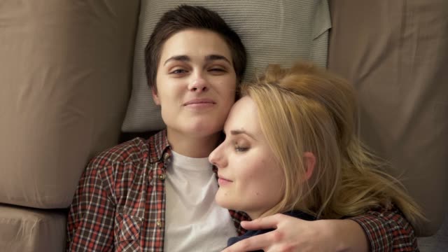 Zwei-junge-lesbische-Mädchen-liegen-auf-der-Couch,-Umarmung,-kuscheln,-schlafen,-Mädchen-mit-kurzen-Haaren-sieht-in-die-Kamera,-LGBT-Familienkonzept,-glücklich,-Top-shot-60-fps
