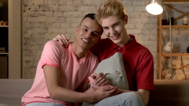 Dos-amigos-homosexuales-multiétnicas-se-sientan-en-el-sofá,-cogidos-de-la-mano,-abrazos,-sonriendo,-mirando-a-la-cámara.-Calidez-de-hogar,-familia,-lgbt-60-fps