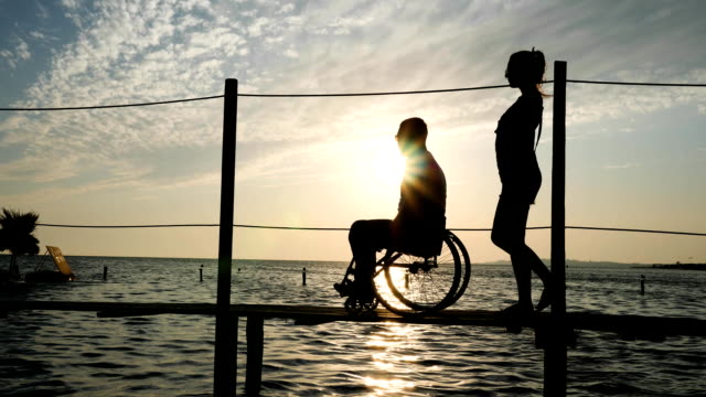 slender-girl-with-man-disabled-on-wheelchair-walking-on-pier-at-sea-against-sky-in-sunset