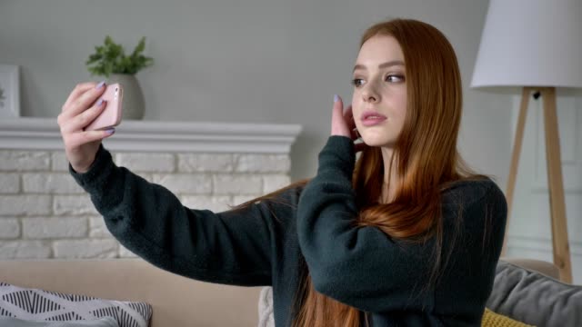Young-beautiful-red-haired-teen-girl-uses-a-smartphone,-makes-selfie,-home-comfort-in-the-background.-60-fps